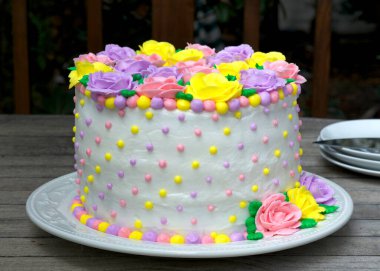 Pastel Gökkuşağı Sarı, Pembe, Mor tereyağı kremalı güller yuvarlak bir pasta üzerinde beyaz kremayla süslenmiş ve ahşap masa üzerinde sarı, pembe ve mor noktalarla süslenmiş.