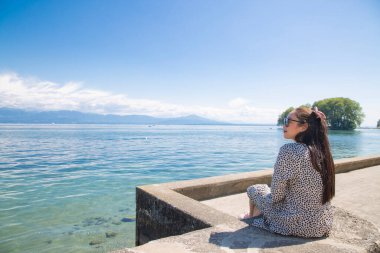 Beyaz tenli, uzun siyah saçlı, siyah gözlüklü güzel bir Asyalı kadın. Gökyüzü açık, turizm ve yaz tatillerinde taşın üzerine oturup göl manzarasını izlemekten mutluluk duyuyorum..