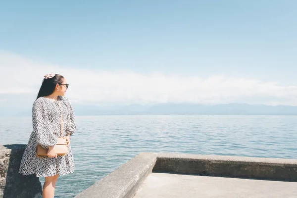 Beyaz tenli, uzun siyah saçlı, siyah gözlüklü güzel bir Asyalı kadın. Gökyüzü açık, turizm ve yaz tatillerinde taşın üzerine oturup göl manzarasını izlemekten mutluluk duyuyorum..