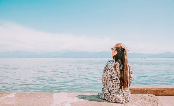Beyaz tenli, uzun siyah saçlı, siyah gözlüklü güzel bir Asyalı kadın. Gökyüzü açık, turizm ve yaz tatillerinde taşın üzerine oturup göl manzarasını izlemekten mutluluk duyuyorum..