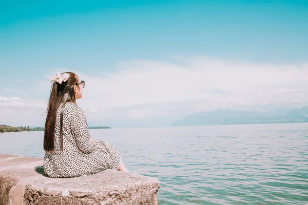Beyaz tenli, uzun siyah saçlı, siyah gözlüklü güzel bir Asyalı kadın. Gökyüzü açık, turizm ve yaz tatillerinde taşın üzerine oturup göl manzarasını izlemekten mutluluk duyuyorum..