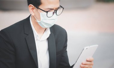 Mikropları, zehirli dumanları ve COVID 19 'u önlemek için doktor maskesi takan Asyalı bir adamın görüntüsü. Tablet ve gazetelerden haberleri okuyor ve dinliyor. Parkta çalışmıyor..
