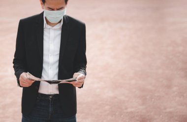 Mikropları, zehirli dumanları ve COVID 19 'u önlemek için doktor maskesi takan Asyalı bir adamın görüntüsü. Tablet ve gazetelerden haberleri okuyor ve dinliyor. Parkta çalışmıyor..