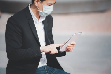 Mikropları, zehirli dumanları ve COVID 19 'u önlemek için doktor maskesi takan Asyalı bir adamın görüntüsü. Tablet ve gazetelerden haberleri okuyor ve dinliyor. Parkta çalışmıyor..