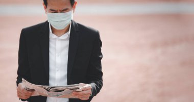 Mikropları, zehirli dumanları ve COVID 19 'u önlemek için doktor maskesi takan Asyalı bir adamın görüntüsü. Tablet ve gazetelerden haberleri okuyor ve dinliyor. Parkta çalışmıyor..