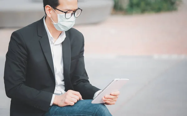 The image face of a Business Asian man wearing a doctor mask to prevent germs, toxic fumes, and COVID 19, He is reading and listening to the news on tablets and newspapers, do not work in the park.