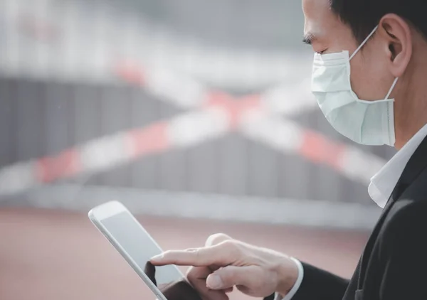 The image face of a Business Asian man wearing a doctor mask to prevent germs, toxic fumes, and COVID 19, He is reading and listening to the news on tablets and newspapers, do not work in the park.