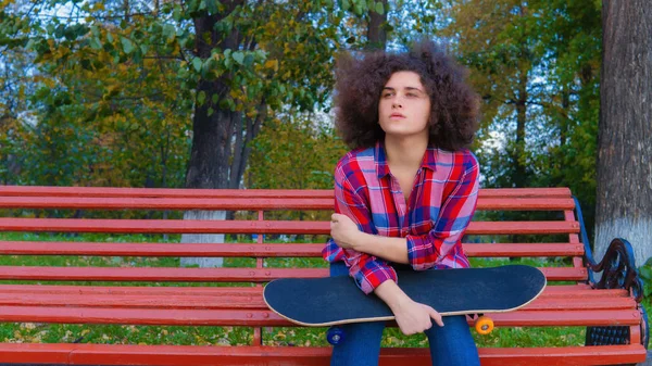 A teenage girl sits with a skateboard in her hands on a wooden park bench. — Stock Photo, Image