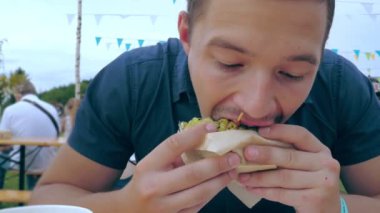 Genç bir adam bir festivalde veya köy panayırında açık hava kafede yeşil çörekle hamburger ya da çizburger yiyor..