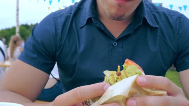 Молодий чоловік їсть гамбургер або чізбургер з зеленою булочкою у кафе під відкритим небом на фестивалі або сільському ярмарку . — стокове відео