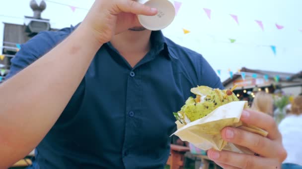 Un joven come una hamburguesa o una hamburguesa con queso con un bollo verde en un café al aire libre en un festival o feria del pueblo . — Vídeos de Stock