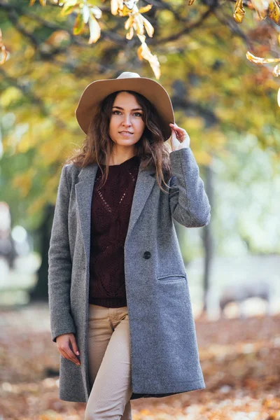 Portrait of beautiful young woman walking outdoors in autumn park in cozy coat and hat. Warm sunny weather. Fall concept. — Stock Photo, Image