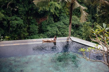 Kenar tropikal sonsuzluk havuzu ormanda üzerinde rahatlatıcı mayo ince seksi esmer kadın. Avuç içi kristal temiz su etrafında. Lüks resort Bali Adası.