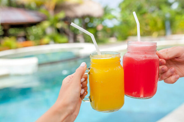 Two colorful fruit shakes in hands. Summer and tropical mood. Cold blended drinks, fruit smoothie. Clink glasses by couple hands