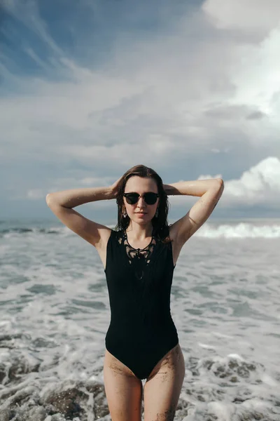 黒水着とサングラスの美しい雲黒砂のビーチでポーズで砂のお尻と美しい少女の背面のファッション撮影 チャングー インドネシア バリ島 — ストック写真