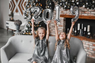 İki mutlu küçük kızlar tutan eller yeni yıl balonlar. Sayı 2019. Noel ağacı ve şömine ile dekore edilmiş oda kanepede oturuyor. Noel zamanı. Yeni yıl kavramı