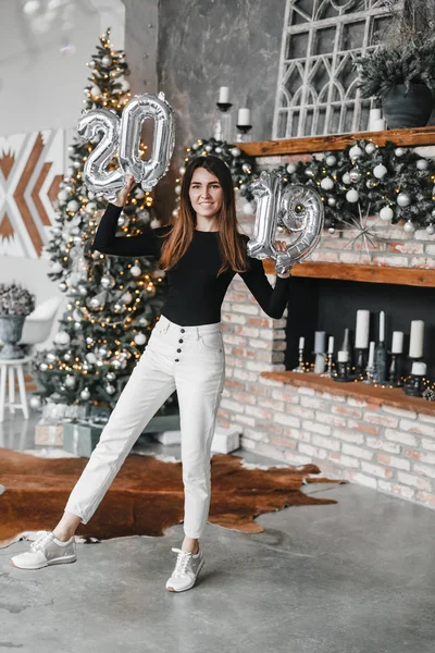 Boldog mosoly lány nő tartja kezében új év lufi karácsonyfa és kandallóval berendezett szobában. Számos 2019. Karácsonykor. Új év-koncepció — Stock Fotó
