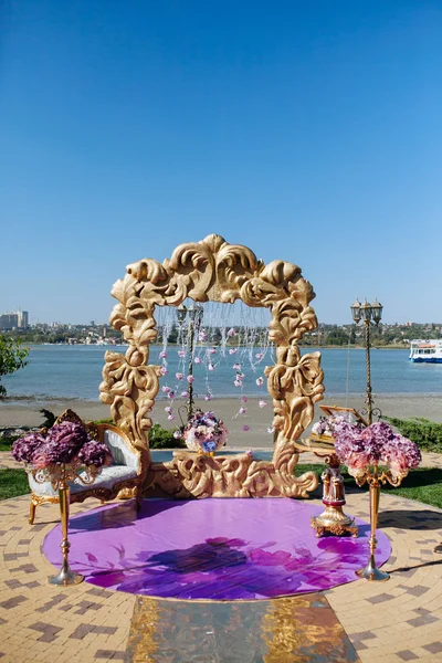 Wedding arch with gold luxury frame and purple flower and chair on riverside. Bride and groom ceremony.