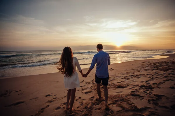 Vista posteriore di ragazza e ragazzo si abbracciano. Silhouette di giovane coppia che cammina sulla spiaggia al tramonto sorprendente che si tiene per mano. Colpo incorniciato orizzontale . — Foto Stock