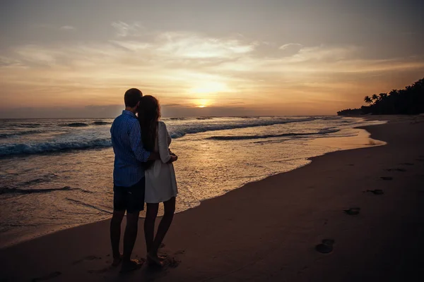 Vista posteriore di ragazza e ragazzo si abbracciano. Giovane coppia godendo il tramonto sulla spiaggia sullo sfondo cielo colorato e riflessione in acqua . — Foto Stock