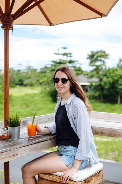 Молодая женщина пьет морковный смузи в баре на открытом воздухе. Брюнетка наслаждается зеленым видом и пьет сок под зонтиком на рисовой террасе . — стоковое фото