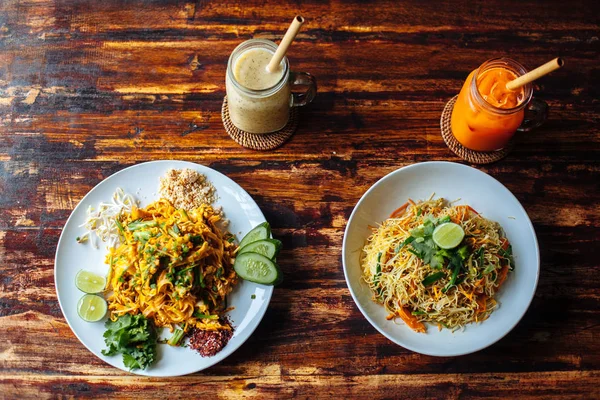 Menú vegetariano saludable Pad Thai, fideos de arroz salteados y fideos singapurenses y batidos de plátano y zanahoria en una mesa de madera.Vista superior — Foto de Stock