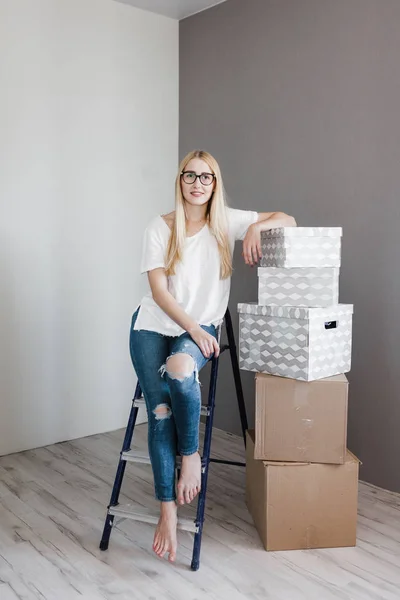 Young woman moving in her new house and doing a home makeover, she is sitting on the ladder with many cardboard boxes around