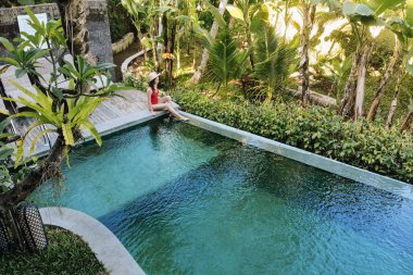 Bali'deki özel havuzda kırmızı bikinili kız, palmiye ağaçlarının güzel manzarasını hayranlıkla izleyebilirsiniz. Lüks tatil. Bali adasında palmiye ağaçlarının güzel bir görünümü ile havuzda dinleniyor Kadın.