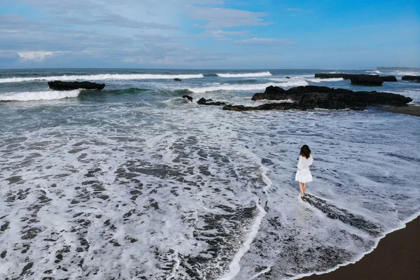 Vista aerea di donna irriconoscibile in abito bianco guardando l'oceano e godersi la giornata estiva sulla spiaggia con sabbia nera. Vacanze a Bali. Foto dal drone. Ricevuto. Foto dal drone — Foto Stock