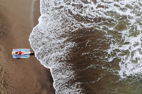 Vista dall'alto della donna in bikini rosso e cappello di paglia abbronzato su telo sulla spiaggia dell'oceano con sabbia nera. Vacanze a Bali. Foto dal drone — Foto Stock