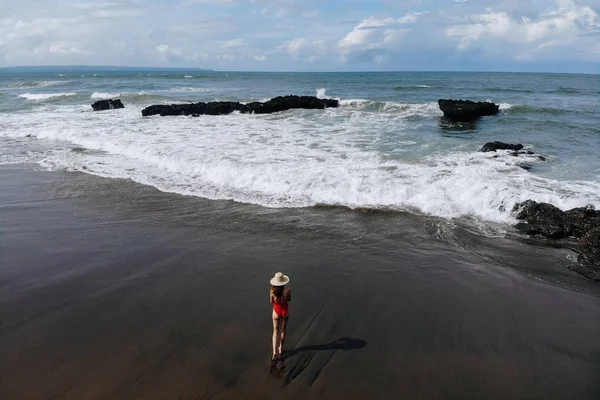 Veduta aerea della donna in bikini rosso e cappello di paglia in mano passeggiando sulla spiaggia dell'oceano con sabbia nera. Vacanze a Bali. Foto dal drone — Foto Stock