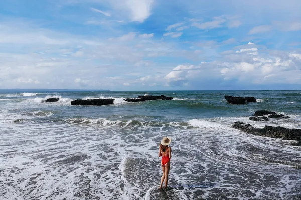 Ragazza in costume da bagno rosso e cappello di paglia godersi la giornata estiva vicino alla spiaggia dell'oceano con sabbia nera. Vacanze a Bali. Foto dal drone. Copia spazio. — Foto Stock