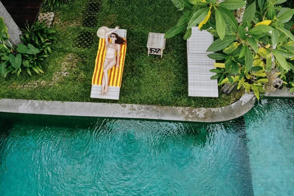 Draufsicht auf eine schlanke junge Frau im beigen Bikini, die sich entspannt und in der Nähe des Luxus-Swimmingpools im Dschungel in Ubud sonnt. Drohnenfoto — Stockfoto