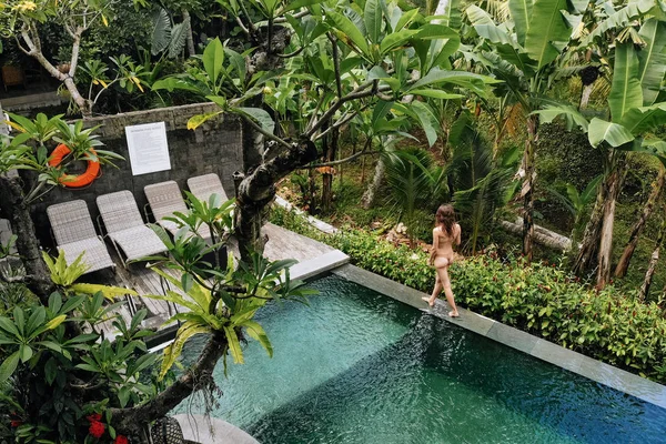 Schlanke sexy brünette Frau im Badeanzug entspannt sich am Rand des tropischen Infinity-Pools im Dschungel. Palmen rundherum und kristallklares Wasser. Luxus-Resort auf der Insel Bali. — Stockfoto