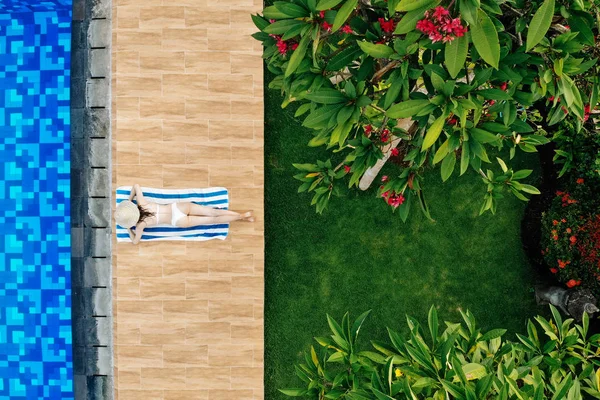 Disfrutando del bronceado. Concepto de vacaciones. Vista superior de la mujer joven delgada en bikini blanco y sombrero de paja tumbado en la toalla cerca de la piscina. Vista trasera, sin rostro —  Fotos de Stock