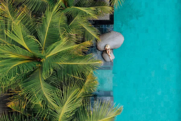 Vista superior de la mujer joven delgada en bikini beige y sombrero de paja relajante cerca de la piscina de lujo y palmeras. Drone foto —  Fotos de Stock