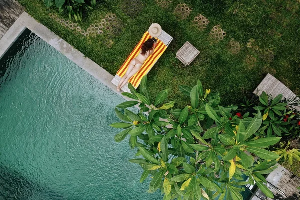 Вид с воздуха на неузнаваемую стройную молодую женщину в бежевом бикини, расслабляющую и загорающую возле роскошного бассейна в зеленом тропике в Убуд.Концепция отдыха. Фотография дрона — стоковое фото