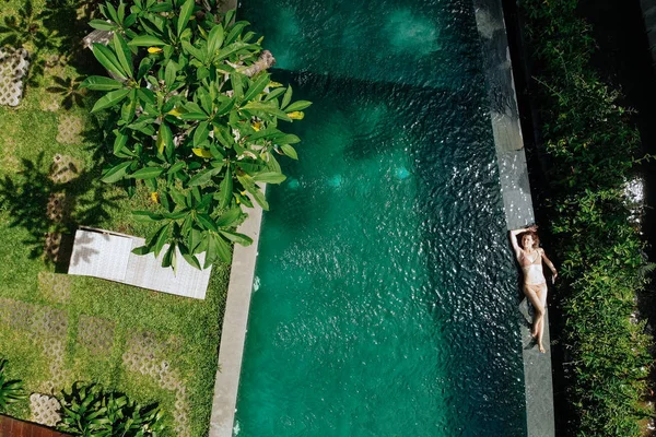 Vista aérea de la mujer en bikini beige relajante y tomar el sol en el borde de la piscina infinita alrededor de las palmeras y la selva. Vacaciones de lujo.Niña descansando en la isla de Bali. Copiar espacio. Concepto de vacaciones —  Fotos de Stock