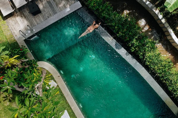 Неузнаваемая женщина в бежевом бикини, расслабляющаяся в бесконечном бассейне на Бали, восхищается прекрасным видом на пальмовые деревья. Принято. Концепция отдыха — стоковое фото