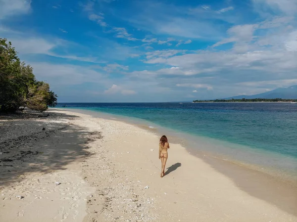 Achteraanzicht van de vrouw in beige bikini wandelen op het strand met Turquoise Oceaan, blauwe lucht. Gili Meno eiland, Bali, Indonesië. Tropische achtergrond en reis concept. Drone Photo — Stockfoto