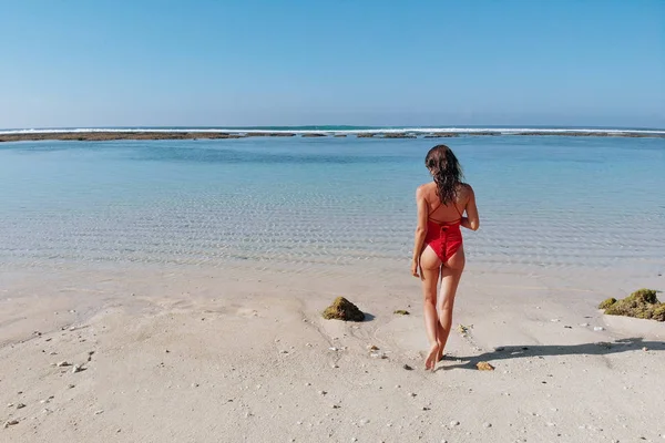 Onherkenbaar vrouw met sexy kont in rode bikini staande op wit zand tropisch strand met blauw helder water en jungle. Bali Indonesia. Tropische achtergrond en reis concept — Stockfoto