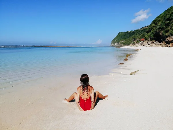 Achteraanzicht van sexy vrouw in rode bikini zittend en poseren op wit zand tropisch strand met blauw helder water en jungle. Bali Indonesia. Tropische achtergrond en reis concept — Stockfoto