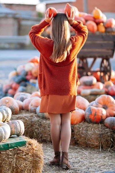 Back view of woman with small pumpkin in hand above head near wooden wagon with pumpkin on farmers market in brown sweater. Cozy autumn vibes Halloween, Thanksgiving day. — Stock Photo, Image