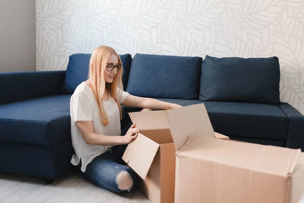 Casa em movimento: Mulher feliz desembalagem caixa em nova casa, menina loira sittng perto de sofá com caixas — Fotografia de Stock