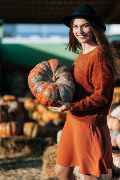 Kobieta w brązowej sukience i swetrze trzyma pomarańczową dynię na tle targu rolniczego. Przytulne jesienne wibracje Halloween party, Święto Dziękczynienia. — Zdjęcie stockowe