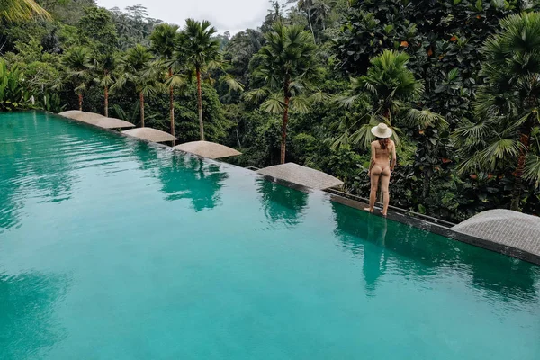 Rückansicht einer Frau, die am Rande des Infinity Pools im beigen Bikini und Strohhut im Dschungel um Bäume spaziert. Urlaubskonzept — Stockfoto