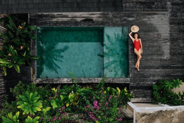 Kırmızı mayo giymiş mutlu bir kadının havuz kenarında çiçeklerle ve yeşilliklerle rahatlarken çekilmiş havadan çekilmiş fotoğrafı, Bali. Tropik arka plan ve seyahat kavramı.