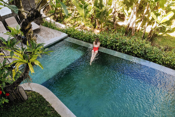 Back view of woman in red bikini relaxing in infinity pool in Bali admires a beautiful view of the palm trees.Luxury holiday.Girl resting on the island of Bali. Copy space. Vacation concept