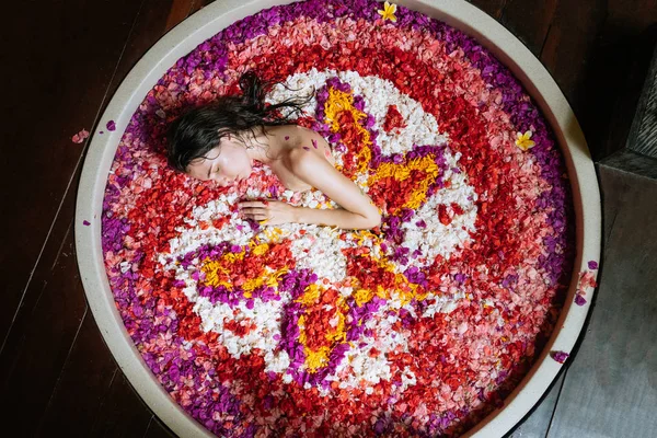 Atractiva Mujer joven en baño con pétalos de flores tropicales y aceites aromáticos. Tratamientos spa para el rejuvenecimiento de la piel. Chica relajante en bañera redonda de lujo —  Fotos de Stock