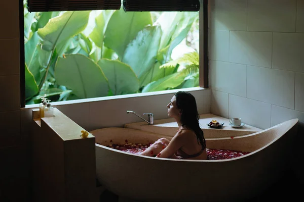 Mujer joven sensible disfrutando en el spa. Bañera de piedra de lujo con vista a la selva cerca de la ventana. Pétalos tropicales orgánicos naturales en el agua. Concepto de tratamiento de belleza . —  Fotos de Stock